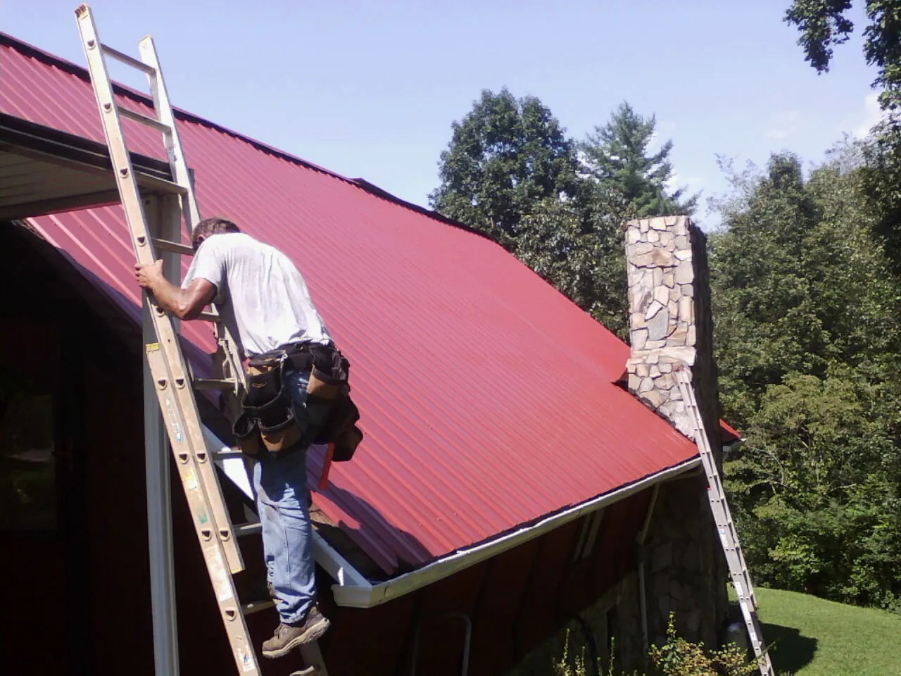 Roofer on a ladder beside a red metal roof and stone chimney — Soffit & Fascia Repair by Roofing Brothers in Tres Pinos